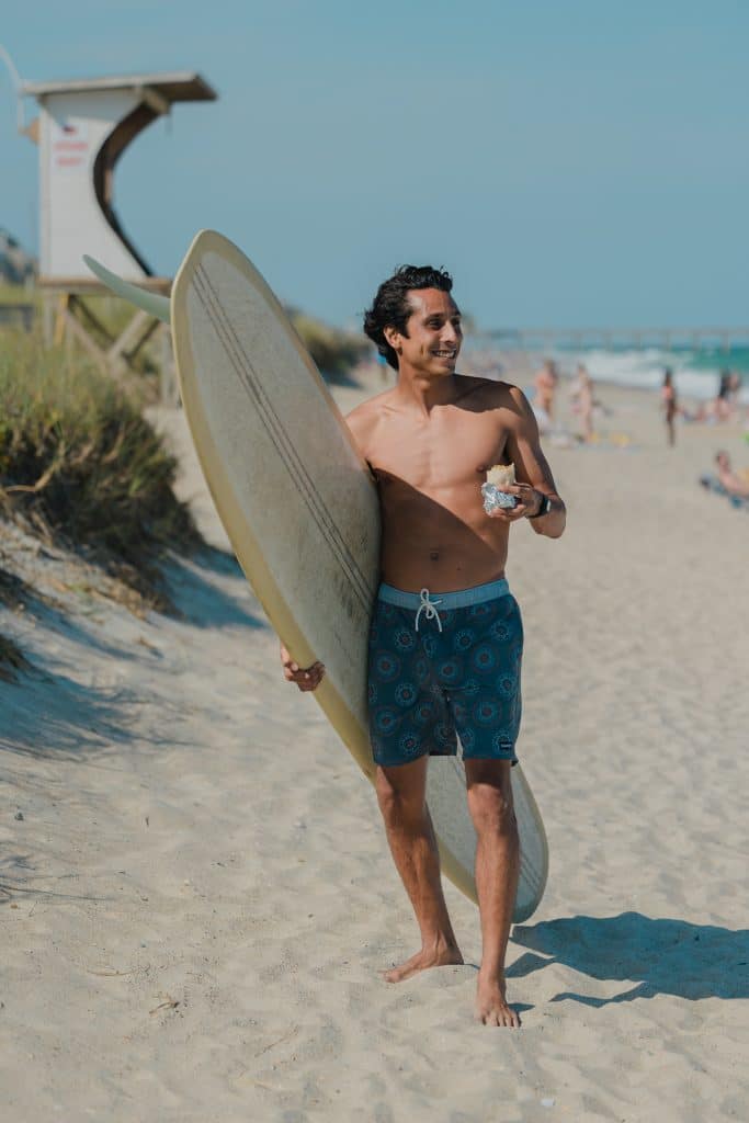 A man in swimming shorts holding a surfboard and a wrap standing on a beach.